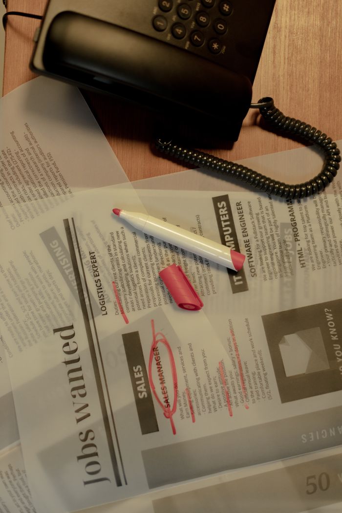 Close-up of a job search scene with a newspaper, red marker, and telephone, indicating job hunting.